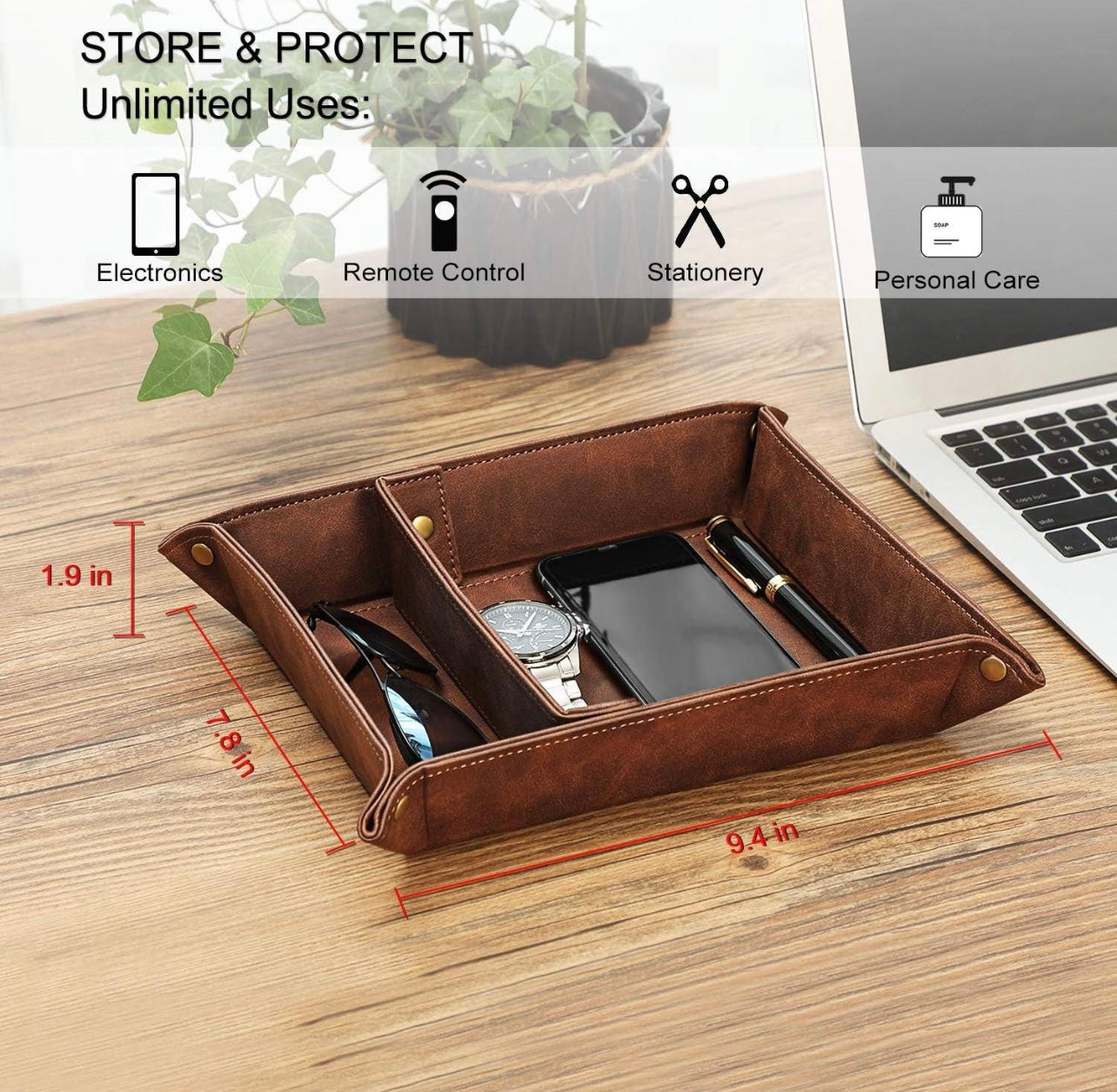 Brown leather tray with compartments on a wooden surface, containing a phone, watch, and pen. Laptop and plant in the background.