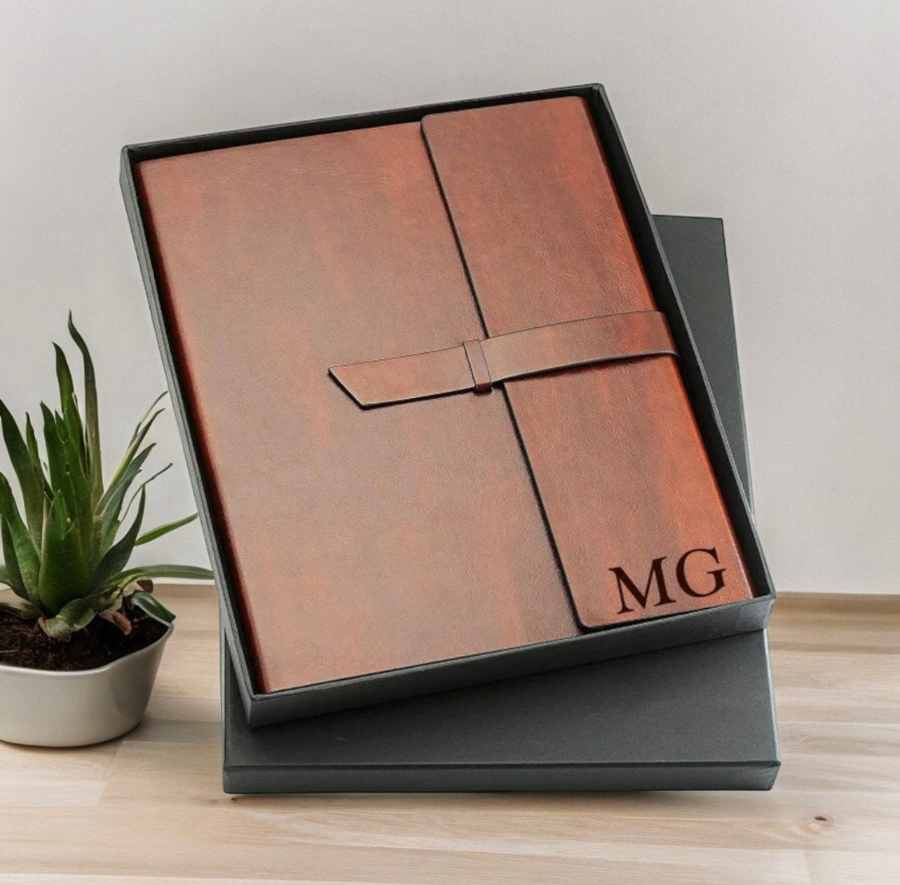 Brown leather-bound notebook with 'MG' monogram in a black box on a light wooden surface.